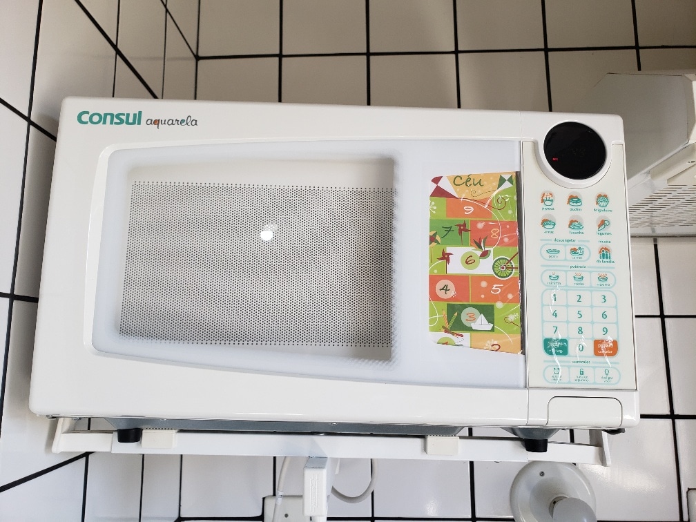 A white microwave is mounted on a wall, featuring a clear window and a colorful control panel with various settings. The front displays a vibrant design with food illustrations, and the microwave is positioned within a tiled kitchen area.