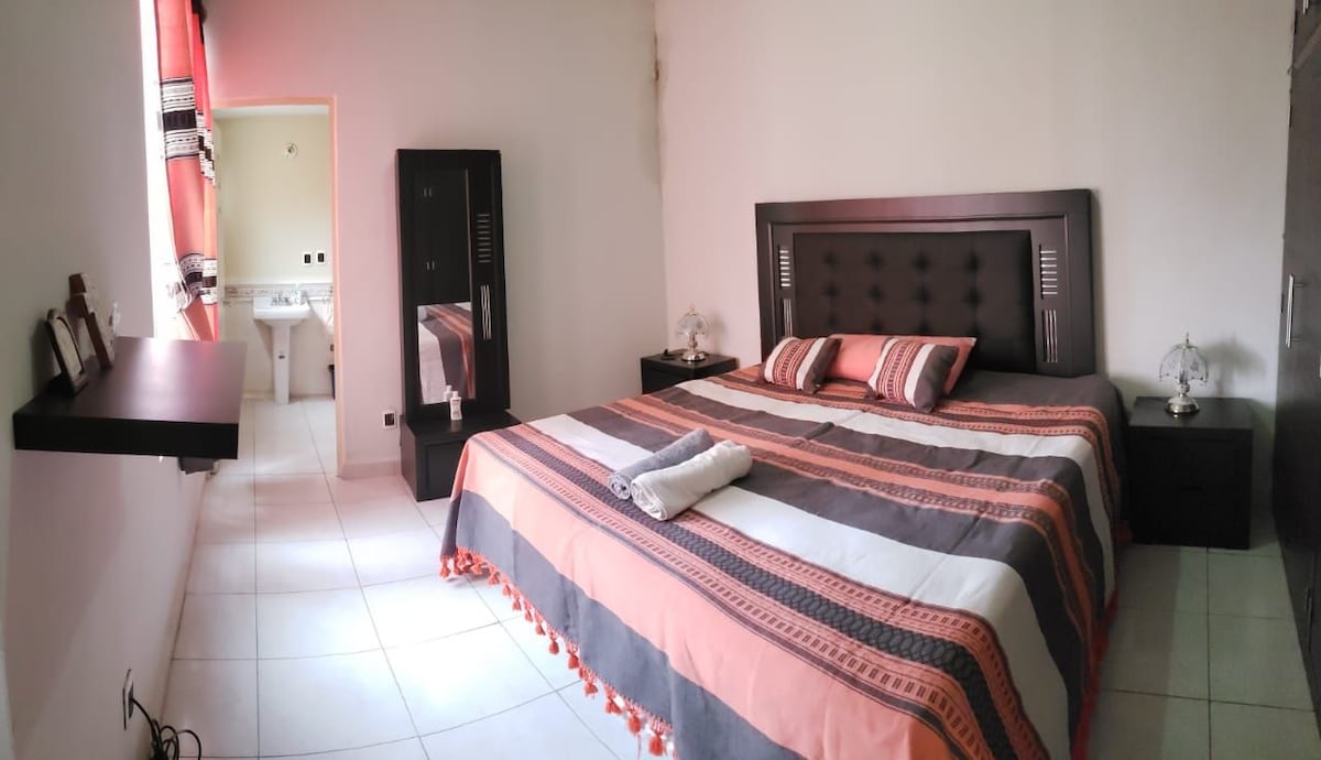 A spacious bedroom features a large bed adorned with colorful, striped bedding. Nightstands are placed on either side of the bed, with lamps providing soft lighting. Bright white tiles cover the floor, and a doorway leads to an adjacent bathroom.