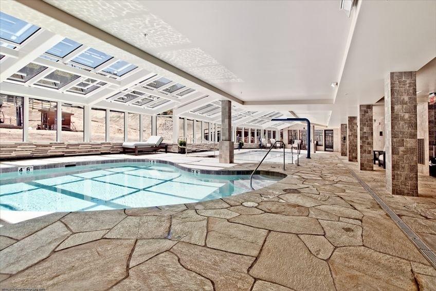 An inviting indoor pool area features a spacious pool surrounded by natural stone flooring. Large skylights allow natural light to illuminate the space, highlighting seating areas along the windows. The atmosphere is tranquil, perfect for relaxation and rejuvenation.