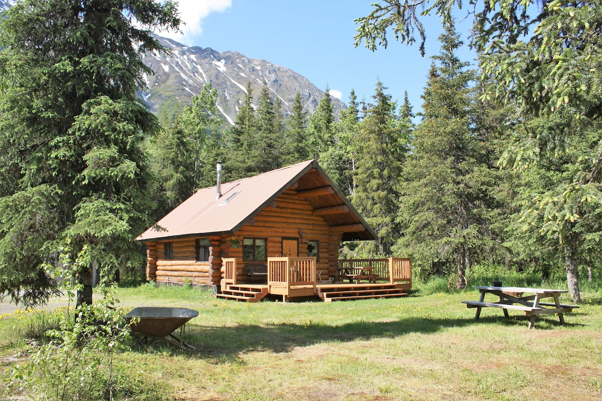 The log cabin is set amidst a wooded landscape, featuring a spacious front deck with a picnic table and lounge chair. Surrounding trees offer shade, while mountains rise in the background, enhancing the serene outdoor environment.