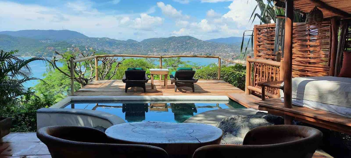 An outdoor terrace presents stunning views of the mountains and ocean. A private dipping pool reflects the sky, while two lounge chairs are positioned beside it. A circular table sits nearby, surrounded by natural greenery, offering a serene space to relax.