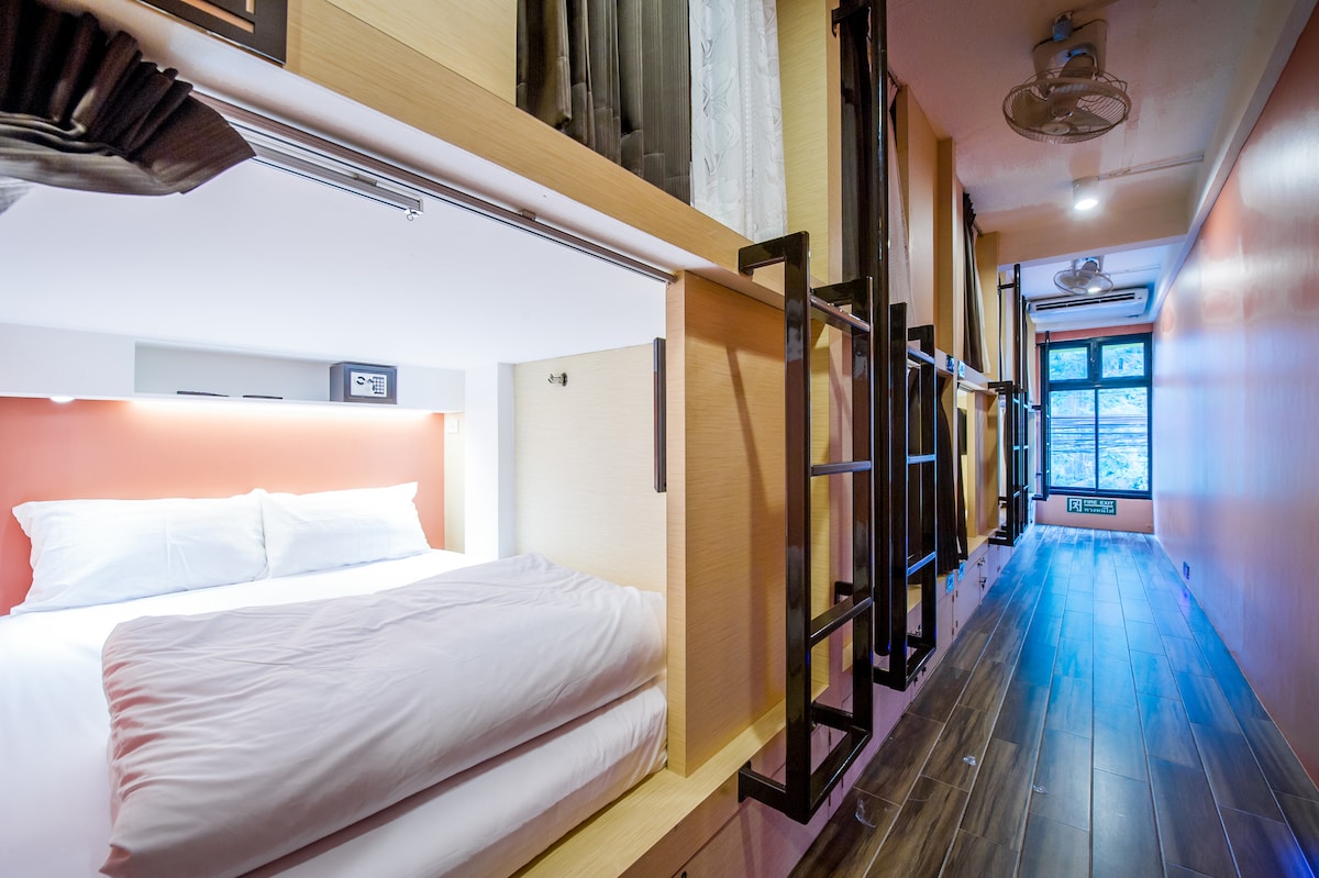 A compact sleeping area is depicted with a spacious bed fitted with crisp white bedding. The warm orange wall enhances the inviting feel. A sleek, wooden floor runs through the room, leading towards a window framed by sheer curtains, allowing natural light to filter in.