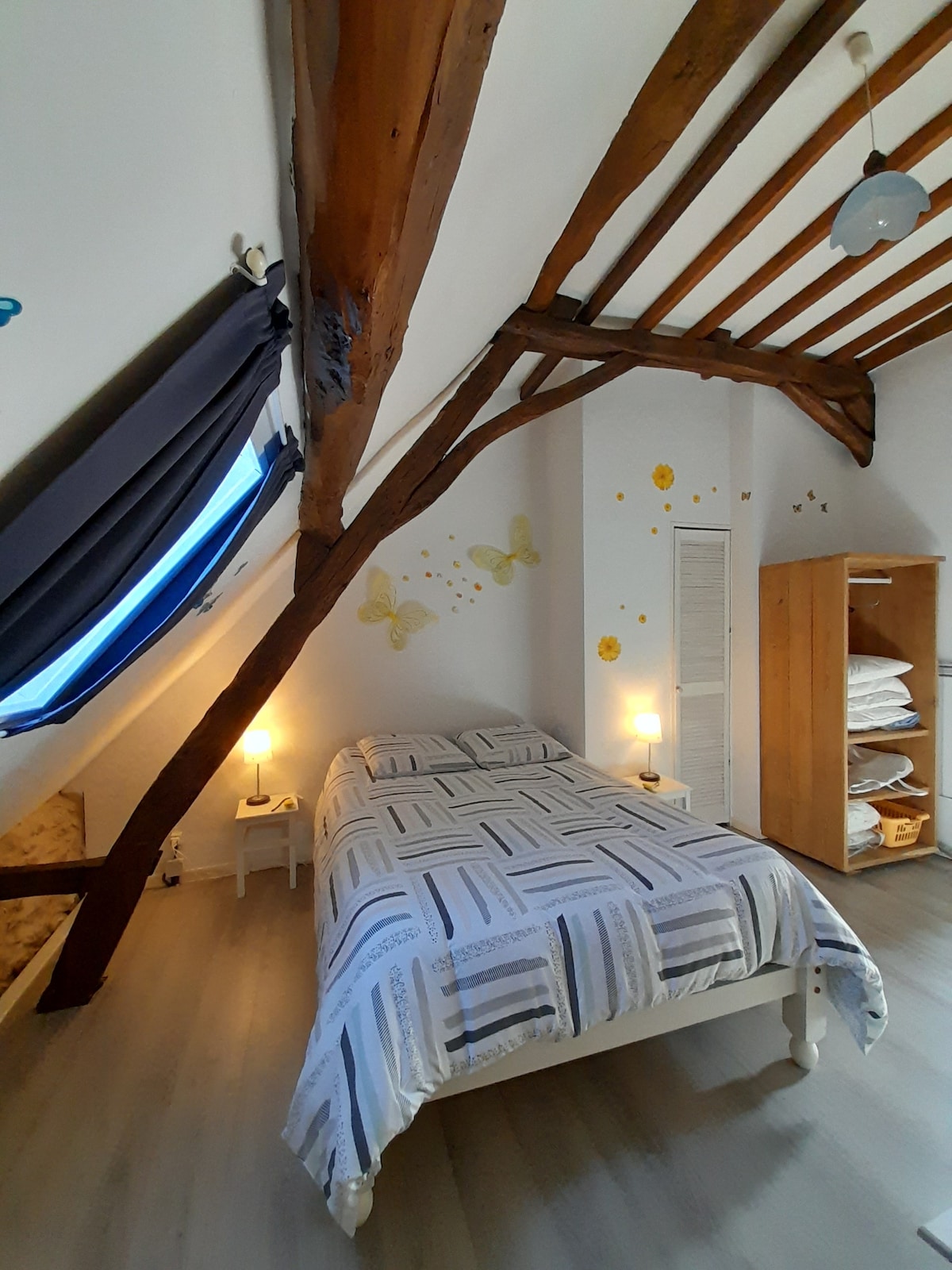 A cozy bedroom features exposed wooden beams and a gently sloped ceiling. A spacious bed is dressed in light linens, flanked by bedside tables with lamps. A wardrobe provides additional storage, while colourful wall art adds a cheerful touch. Natural light filters through a window with draped curtains.