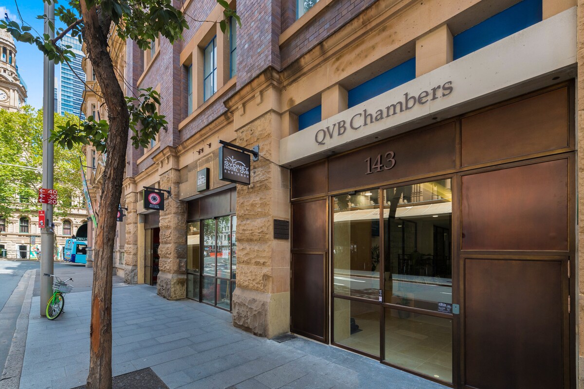The exterior of the building is showcased, featuring a modern facade with large glass windows framed by a bronze entrance. The street is lined with trees and pedestrians, highlighting the urban environment with nearby shops and structures visible in the background.