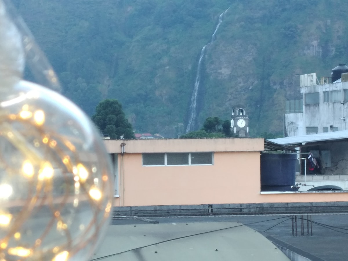 A distant waterfall cascades down a green mountain, visible beyond a clock tower. A portion of an adjacent building is seen in the foreground, featuring a flat roof and windows. Decorative round lights are softly illuminated in the lower left corner.