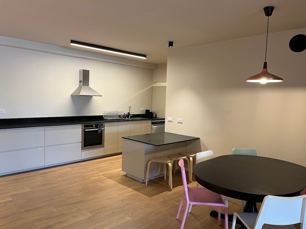 A modern kitchen and dining area feature sleek cabinetry and a spacious island with seating. Stainless steel appliances and a minimalist design create a functional cooking space. A round dining table with colorful chairs adds a touch of vibrancy, complemented by warm lighting from the pendant fixture.