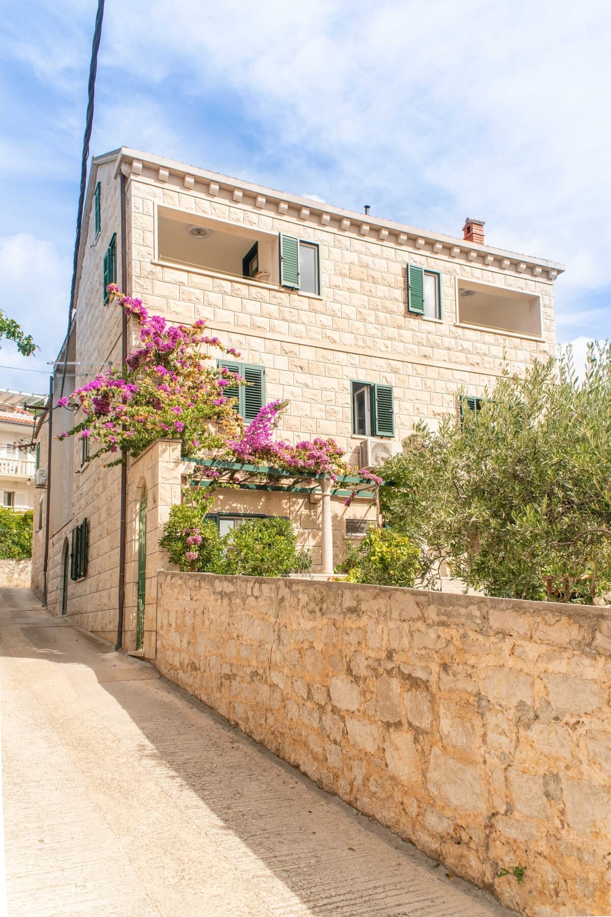 A charming stone house with green shutters and a balcony, adorned with vibrant purple flowers and surrounded by greenery. The structure features a welcoming entrance and is set on a quiet street, showcasing the peaceful ambiance of the neighborhood.