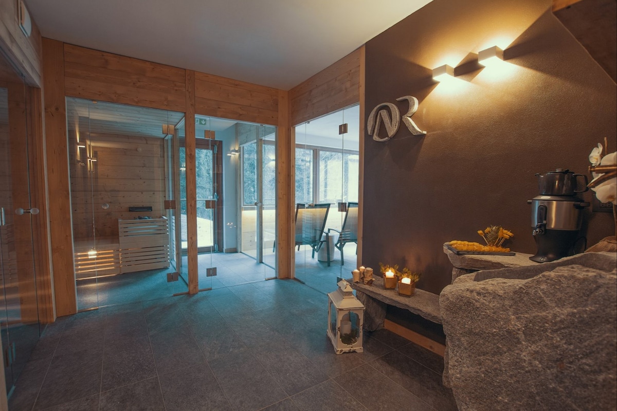 A serene wellness area is seen with large glass walls allowing natural light to filter in. Wooden details enhance the ambiance, while a stone bench is positioned beside an array of candles and a coffee machine. A sauna entrance is visible in the background.