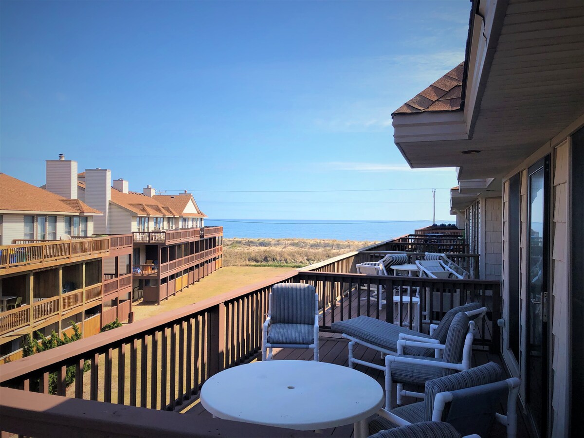 A spacious balcony is equipped with comfortable seating and a small round table, overlooking the ocean and golden sandy beach. The view includes nearby buildings and a clear blue sky, providing a serene atmosphere for relaxation.