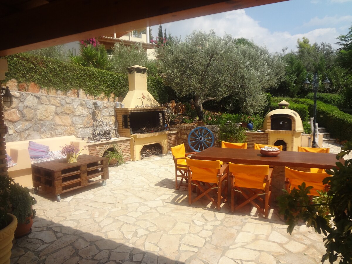 An outdoor barbecue area featuring a stone fireplace and an outdoor oven is surrounded by greenery. A large wooden table with orange chairs accommodates guests. The stone-paved space offers a sunny and inviting setting, complemented by a decorative blue wheel and lush olive trees.