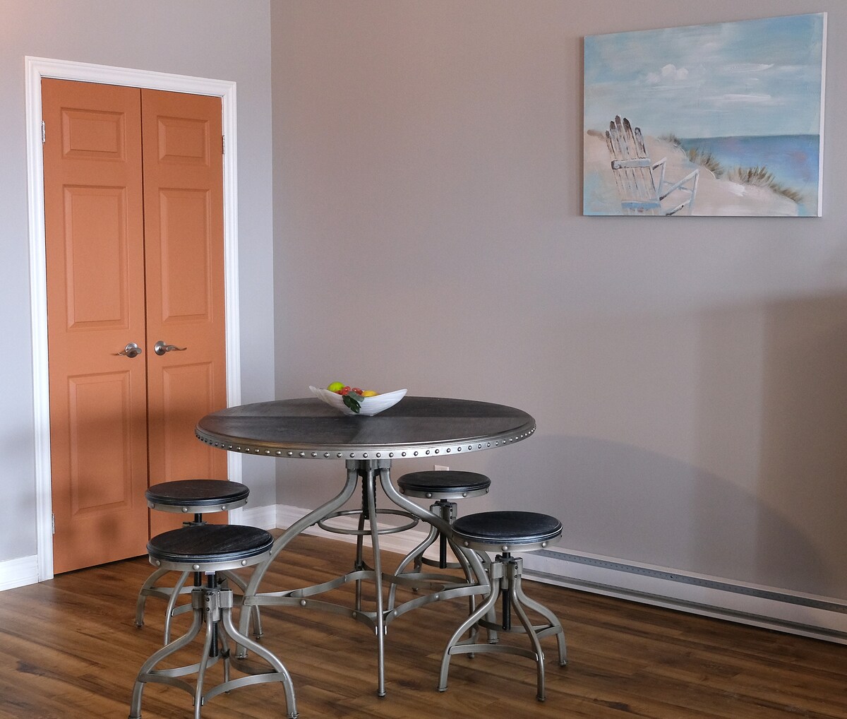 A round dining table is surrounded by six stools, positioned against a neutral wall. Two orange doors provide contrast to the space, and a painting depicting beach chairs overlooks the setup. Warm wooden flooring extends throughout the area.