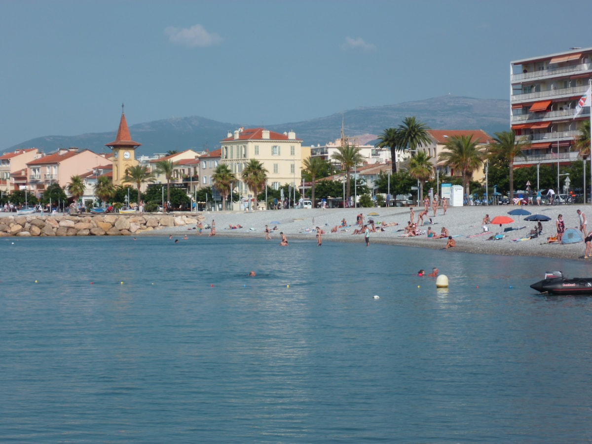 A sandy beach is shown with a calm blue sea reflecting the sky. People are depicted enjoying the shoreline, some in the water and others on the sand under colorful umbrellas. In the background, residential buildings and palm trees create a leisurely atmosphere, complemented by a clock tower.