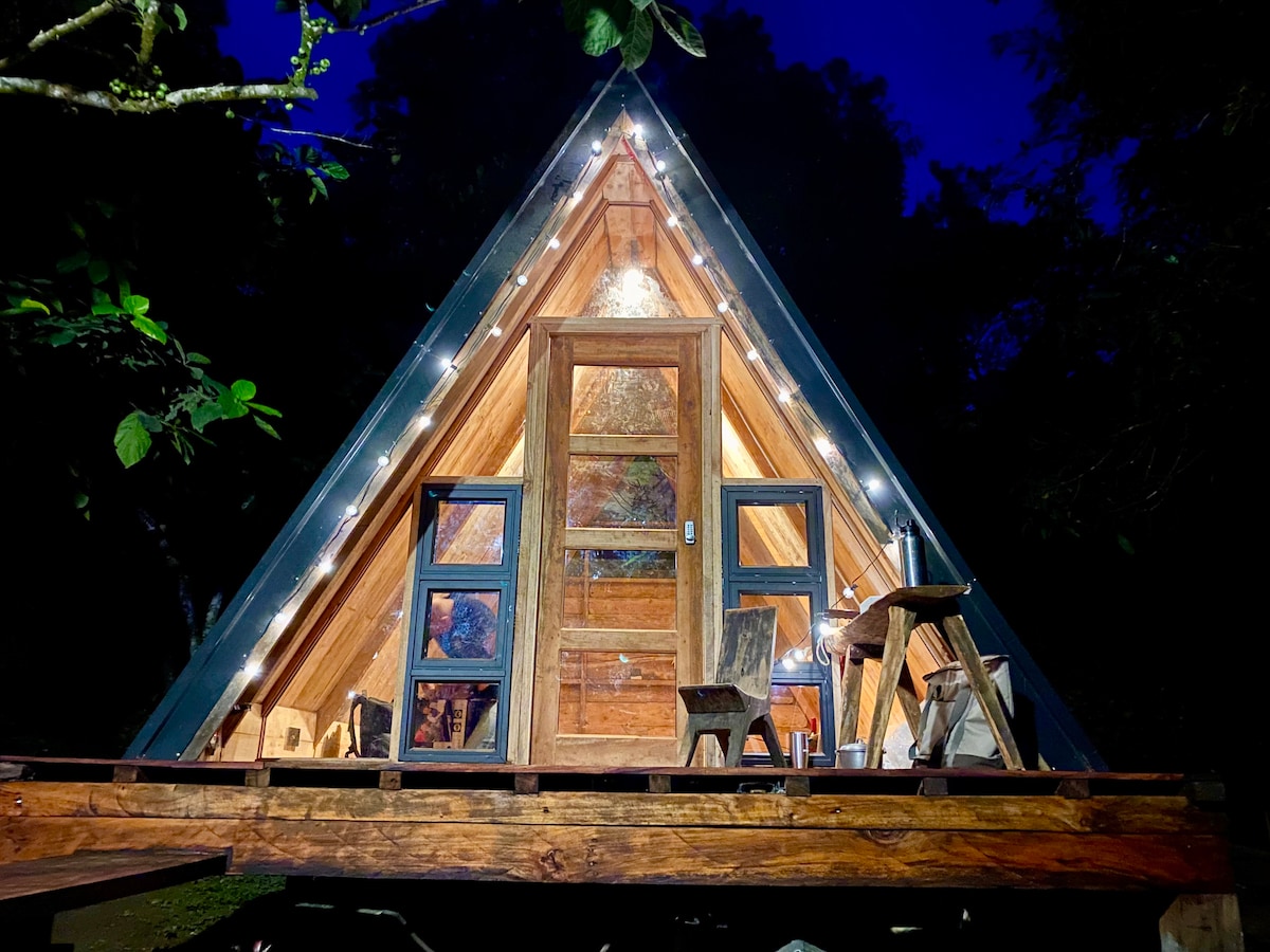 An A-frame cabin is illuminated at night, showcasing its wooden structure and large front windows. String lights frame the entrance, enhancing the natural surroundings. Two rustic chairs are positioned on the deck, inviting relaxation in a serene outdoor setting.