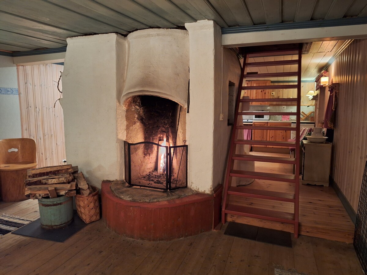 A rustic fireplace is set in a whitewashed stone hearth, with a glowing fire visible inside. Wooden logs are neatly stacked beside it. A wooden staircase leads to the upper level, while warm wood paneling and cabinetry are seen in the background.