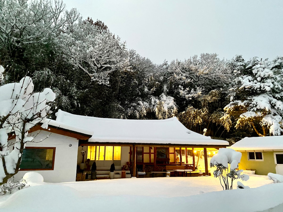A traditional Korean hanok is visible under a blanket of fresh snow, surrounded by snow-covered trees. Soft yellow light glows from the windows, creating a warm contrast against the wintry landscape. The structure features a gently sloping roof and a welcoming porch area.