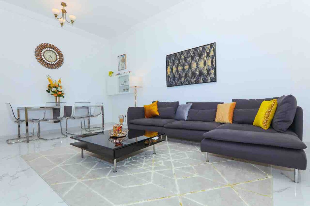 A modern living area features a spacious gray sectional sofa adorned with decorative cushions. A sleek coffee table sits on a patterned rug, while a glass dining table with metallic chairs is positioned next to a wall adorned with a large circular clock and artwork.