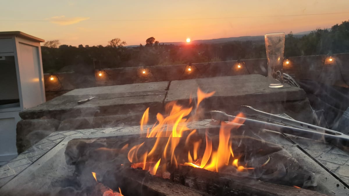 A fire pit filled with glowing flames is shown in the foreground, with the sun setting in the distance. Soft lights surround the area, creating a serene evening ambiance, while the landscape fades gently into the horizon.