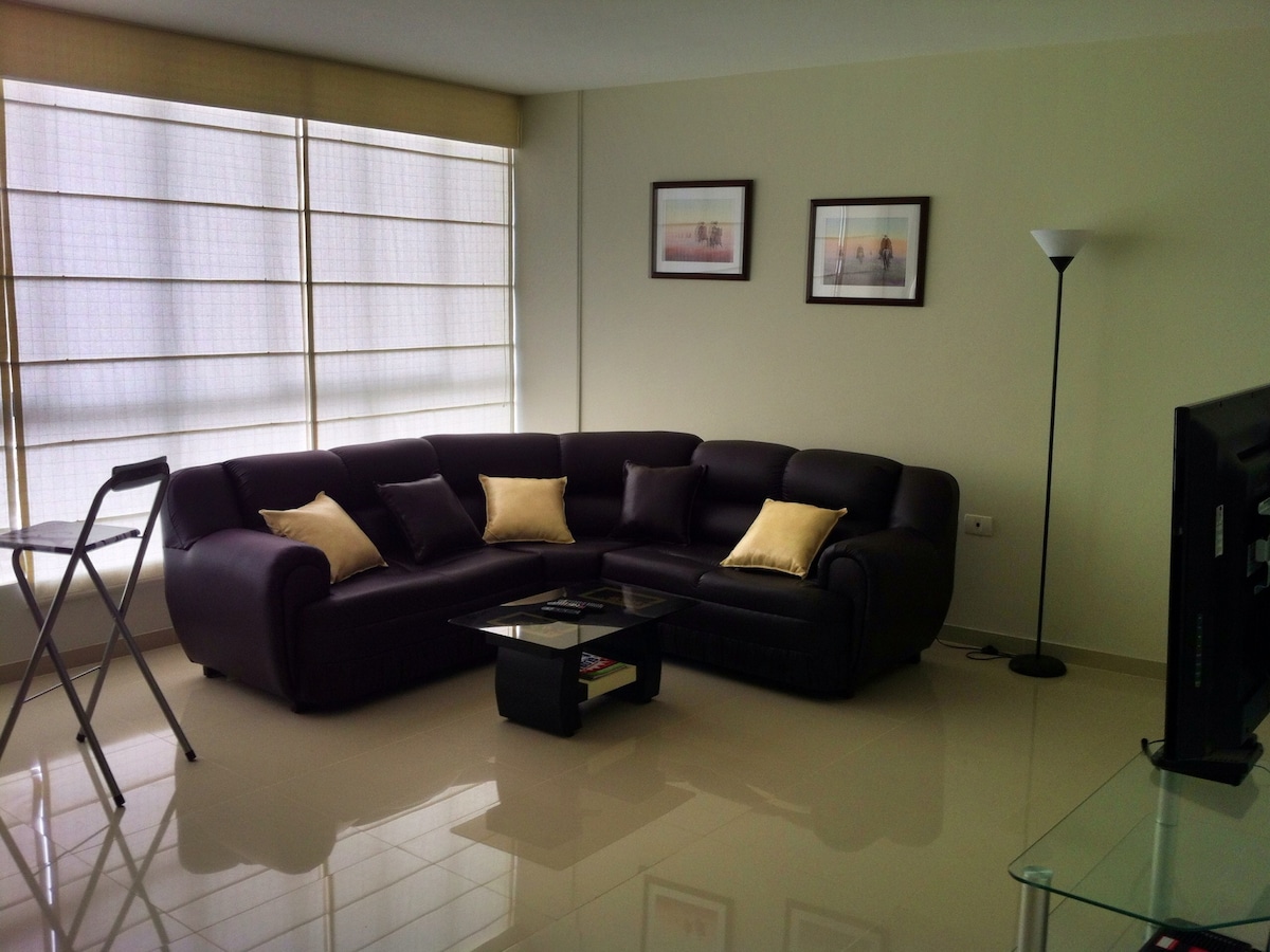 A modern living area features a dark sectional sofa adorned with decorative pillows. A glass coffee table centers the space, accompanied by a simple floor lamp. Natural light filters through large windows, enhanced by light-colored blinds, illuminating the light tile flooring.