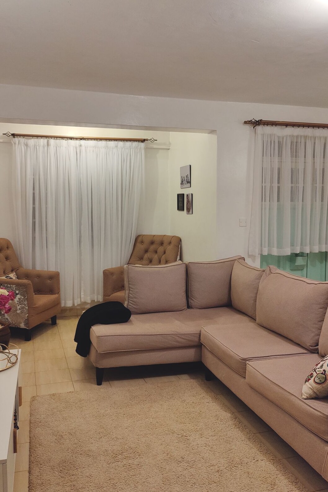 A light-filled living area features a corner sectional sofa in soft beige fabric, accompanied by two textured armchairs. Sheer white curtains adorn the windows, allowing natural light to brighten the space. A plush area rug adds warmth beneath the seating arrangement.