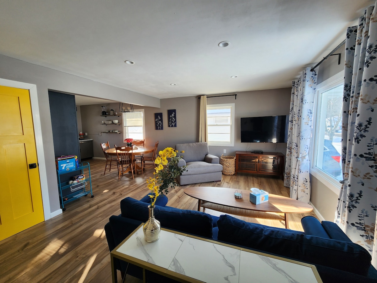 A bright living area features a navy blue sofa with a decorative vase of yellow flowers on a coffee table. A wooden dining table is set in the background, accompanied by chairs. Two large windows allow natural light to fill the space.