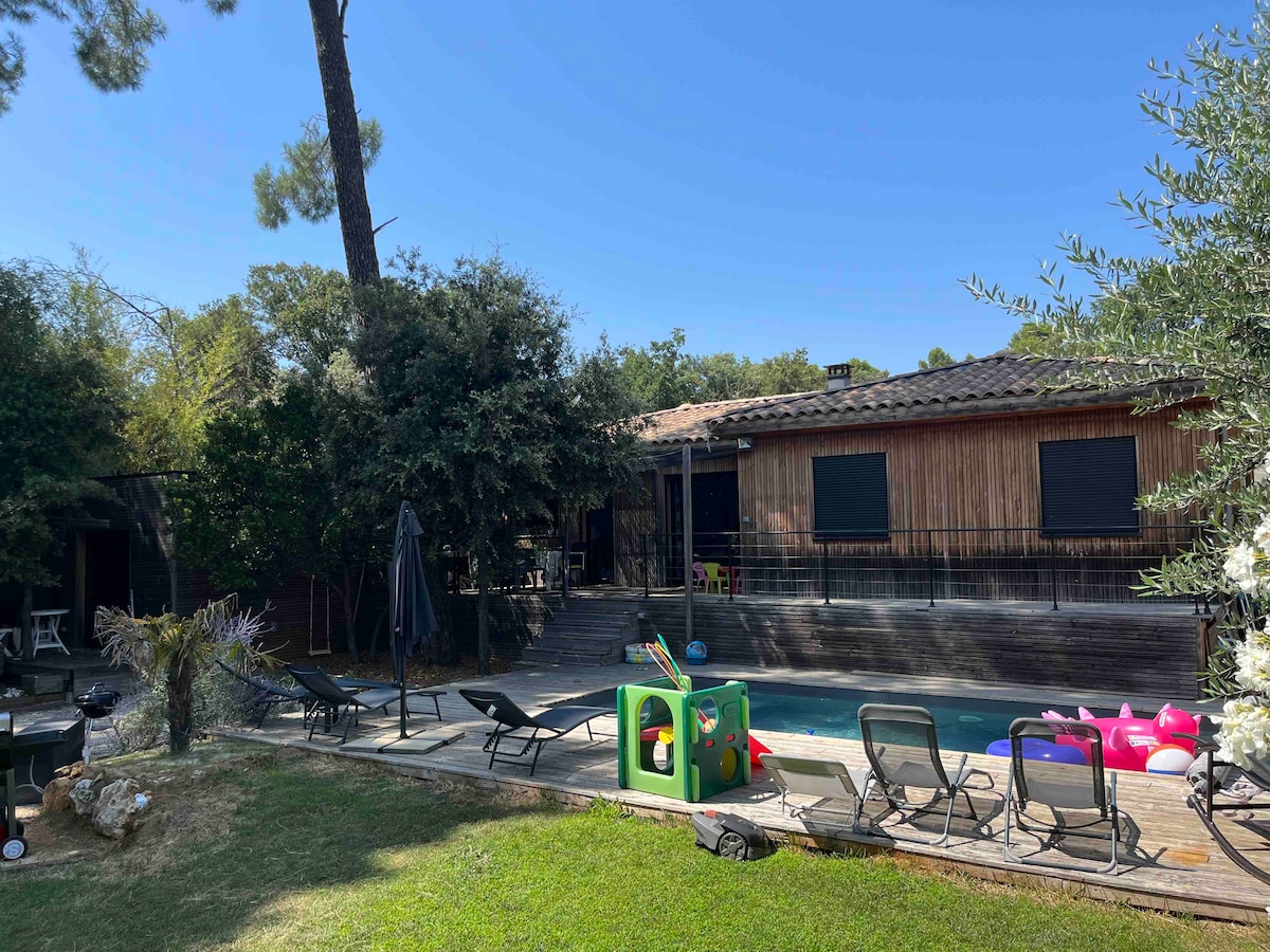 The backyard features a refreshing swimming pool surrounded by loungers and colorful inflatable toys. A wooden deck leads to the house, framed by lush greenery. An umbrella provides shade, while a children’s play area is set nearby, inviting leisure and relaxation.
