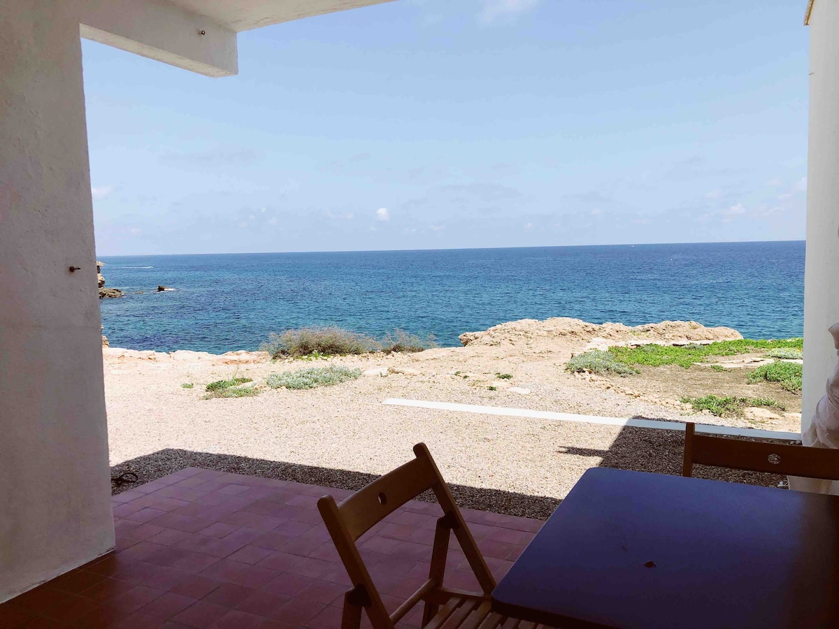 An outdoor terrace offers a view of the calm sea and coastline. A simple wooden table and chairs are positioned in the foreground, allowing for relaxation while enjoying the coastal scenery. The horizon meets a clear blue sky, enhancing the tranquil environment.
