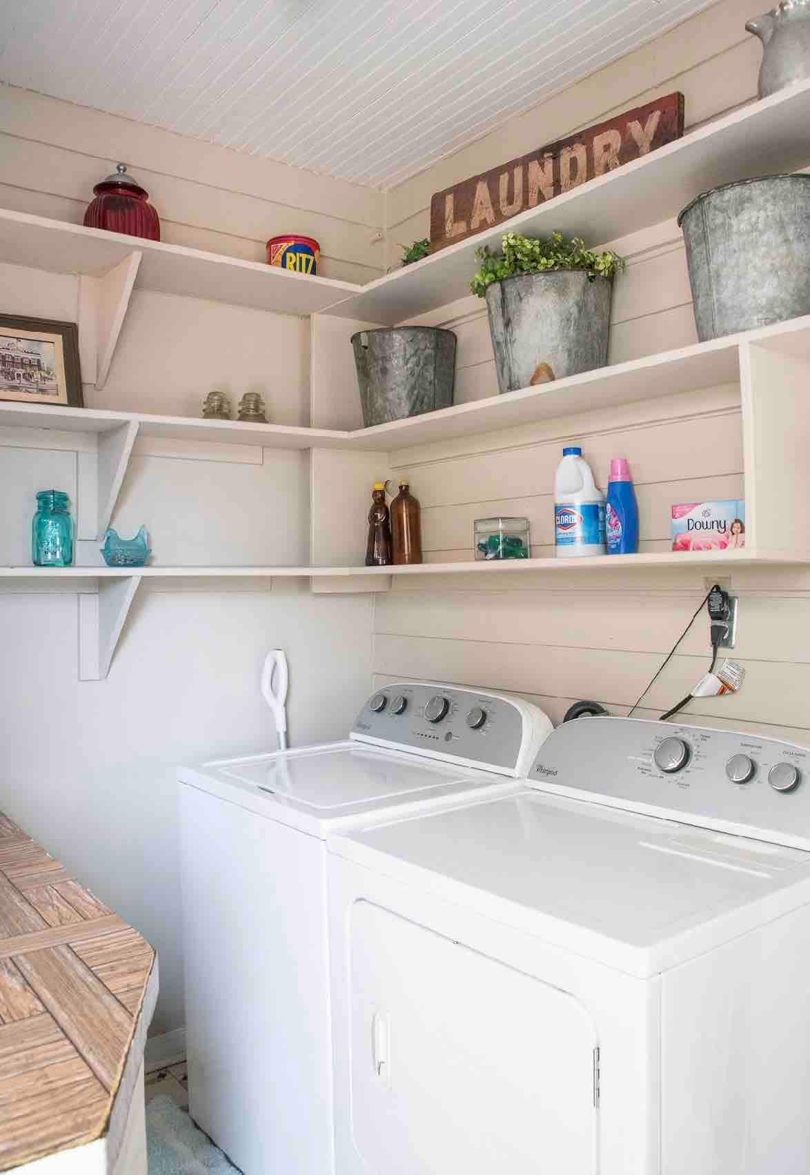 A clean laundry room is shown featuring a washer and dryer side by side. Above, shelves are lined with decorative storage bins and bottles. A 'LAUNDRY' sign is mounted on the wall, and various items enhance the organized space.