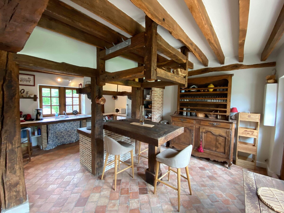 The kitchen displays exposed wooden beams and earthy tile flooring. A rustic wooden table is accompanied by high-backed light-colored chairs. A wooden cabinet holds dishware, while natural light floods the space through multiple windows, creating a warm, inviting cooking area.