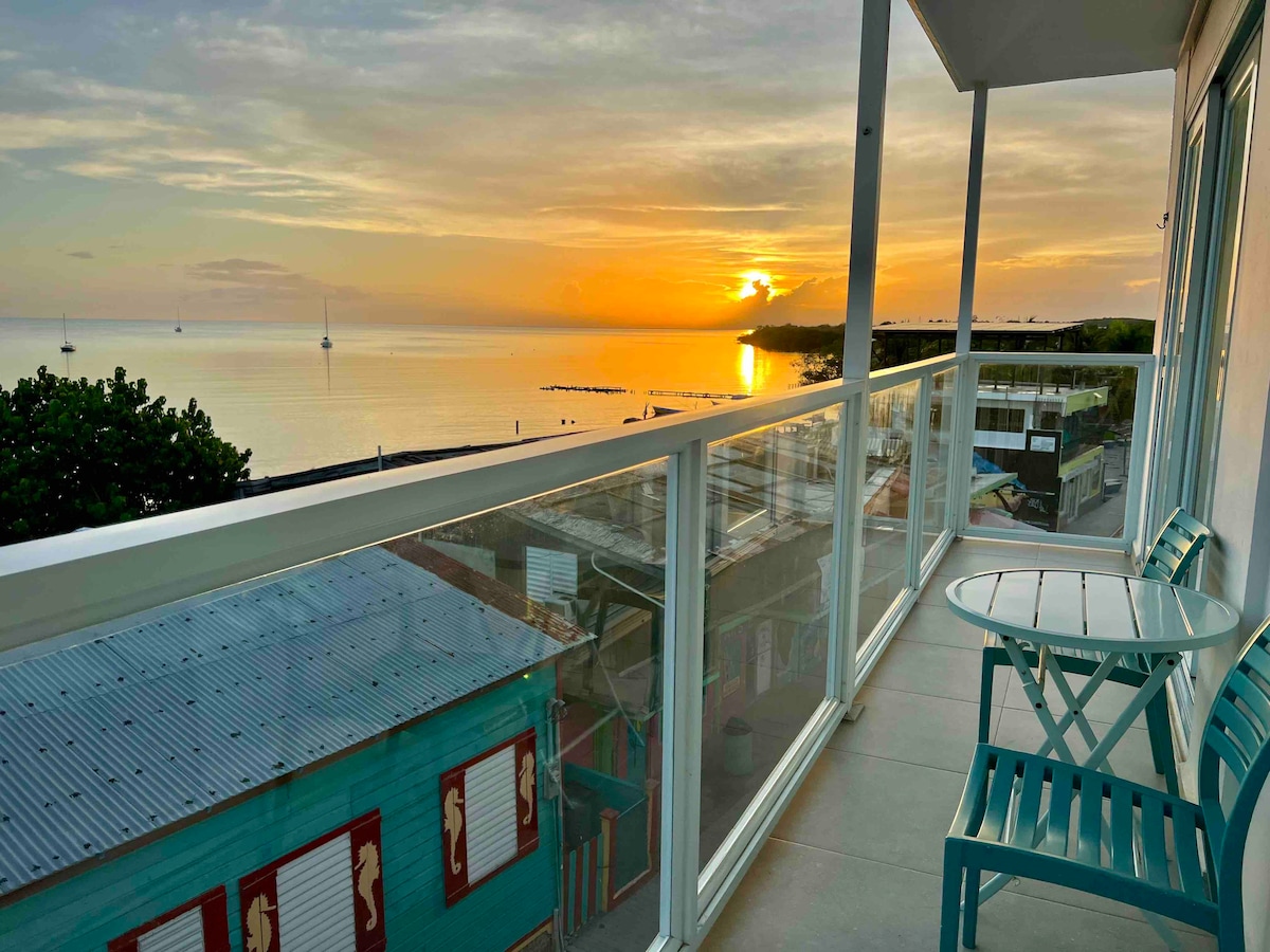 A spacious balcony is shown with a table and chairs, offering a scenic view of the ocean at sunset. The vibrant colors of the sky reflect on the water, while boats are visible on the horizon. Colorful buildings line the street below.