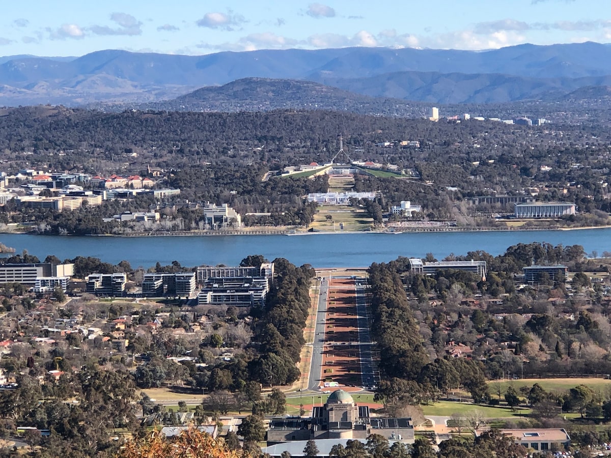A wide view of Canberra showcases the tree-lined avenues leading towards Parliament House, with the clear waters of Lake Burley Griffin visible in the foreground. The distant mountains add depth to the serene landscape, enhancing the connection between nature and the city.