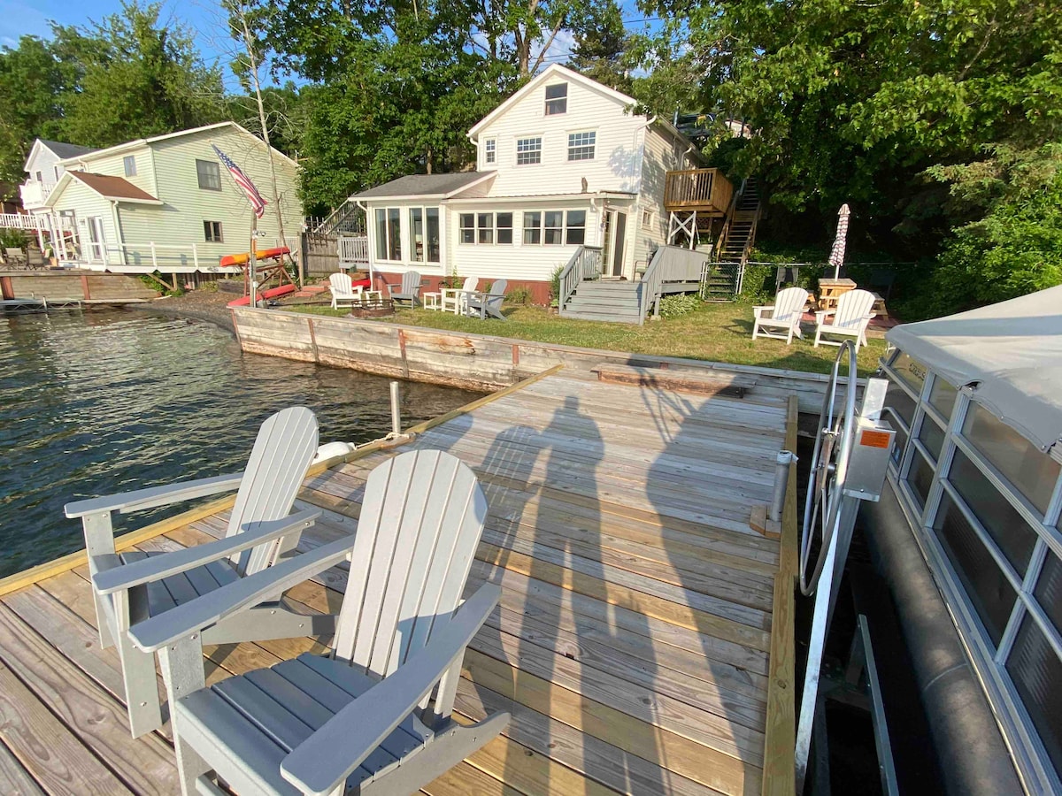 A wooden dock extends into the water, featuring two Adirondack chairs positioned for relaxation. The cottage, with multiple stories and a welcoming entryway, is visible in the background, surrounded by trees and adjacent buildings. Sunlight illuminates the scene, highlighting the serene lakeside setting.