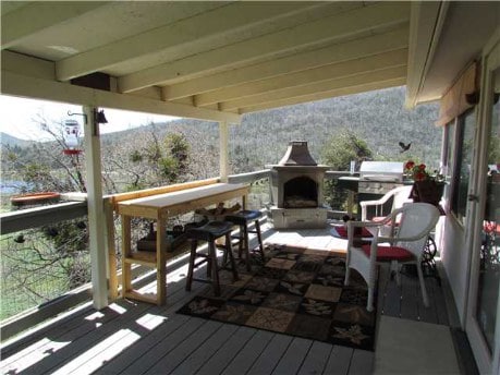 A spacious porch features a wooden table with stools, a gas fireplace, and white chairs. A patterned area rug adds texture to the space, while nearby trees and mountains can be seen in the background. Bird feeders hang nearby, inviting wildlife observation.