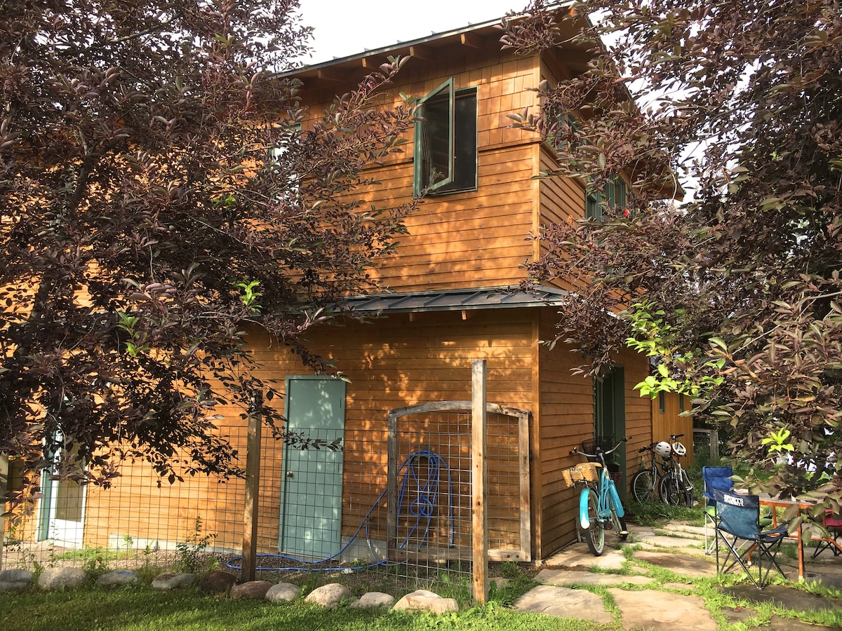 The exterior of a wooden multi-story house is surrounded by lush foliage and vibrant trees. A fenced area is visible in the foreground, providing a safe space for pets. Bicycles are parked nearby, along with a small patio space featuring natural stone pathways.