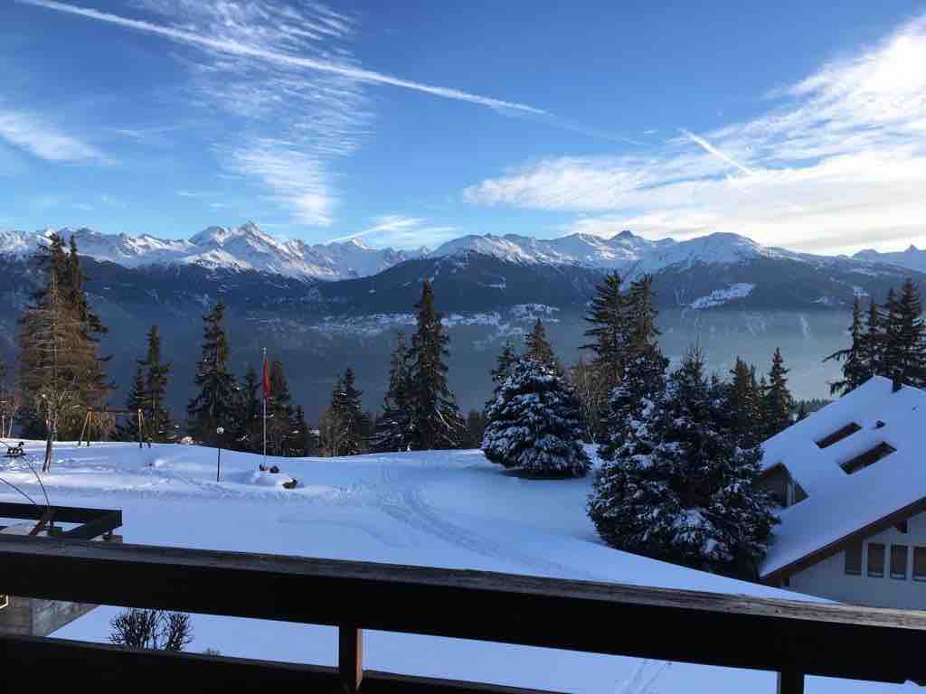A snow-covered landscape unfolds from the balcony, featuring majestic mountain peaks under a clear blue sky. Evergreen trees dot the area, and a gentle layer of snow blankets the ground, enhancing the serene winter atmosphere.