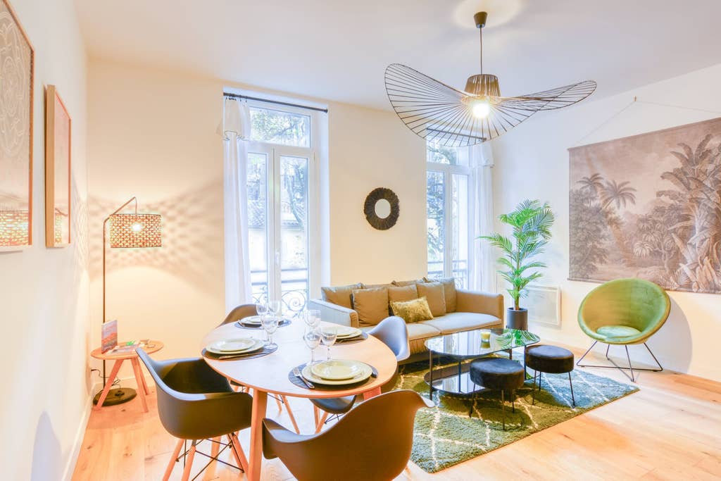 A modern living area is arranged with a circular dining table set for four and a cozy sofa. Large windows invite natural light, with a stylish pendant light overhead. A vibrant green armchair adds a pop of color, complemented by plants and decorative wall art.