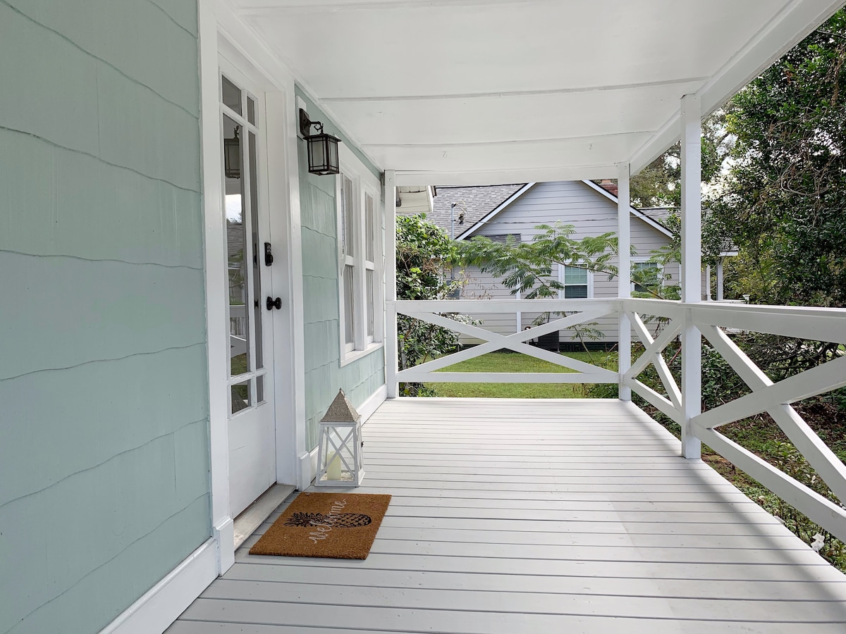 A spacious front porch features light blue walls and white railing, inviting relaxation. A lantern hangs by the entrance door, and a welcome mat is positioned on the weathered wood floor. Lush greenery is visible in the background, enhancing the serene outdoor space.