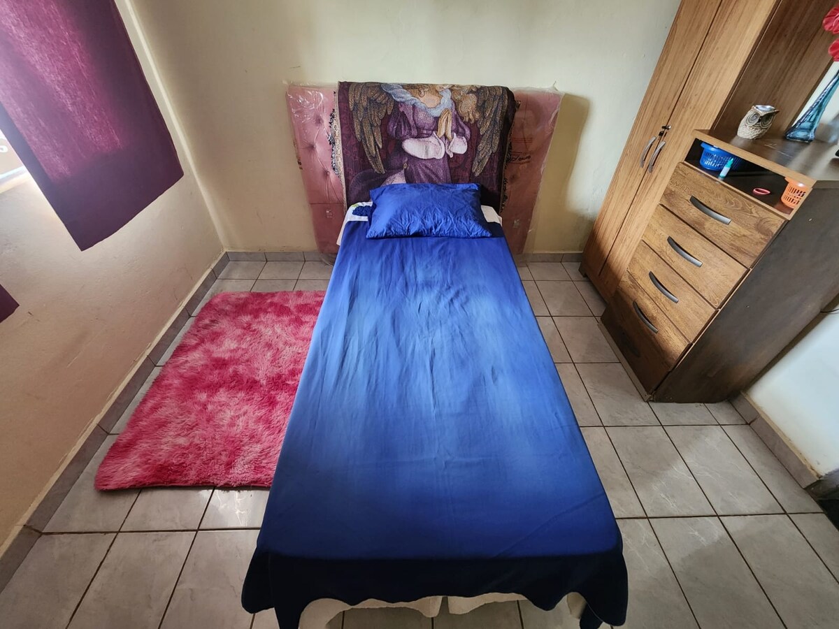 A neatly arranged sleeping area features a twin bed with a blue bedspread and a decorative headboard displaying a figure. A soft pink rug lies beside the bed, and a wooden dresser is positioned against the wall, providing storage space.