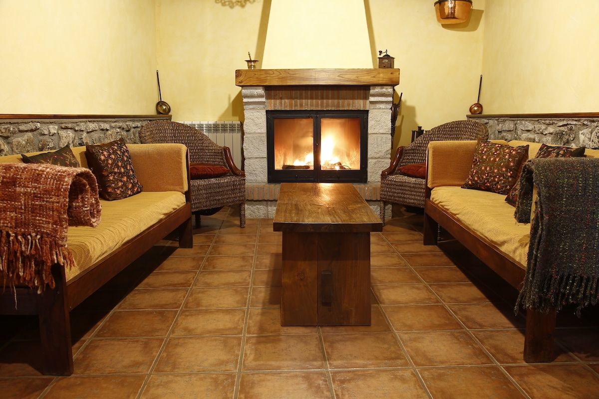 A welcoming living area is presented with two comfortable sofas adorned with patterned cushions, flanking a wooden coffee table. A stone fireplace is centrally located, offering a warm glow, while beige walls complement the rustic tiled flooring.