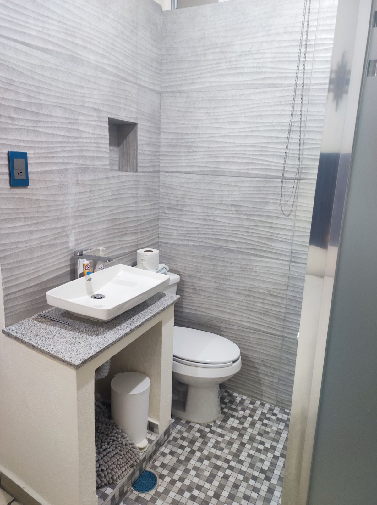 A compact bathroom features a modern white sink atop a gray countertop. The toilet is positioned beside the sink. Textured gray walls and a patterned floor add visual interest, while essential toiletries, including tissue and soap, are neatly arranged.
