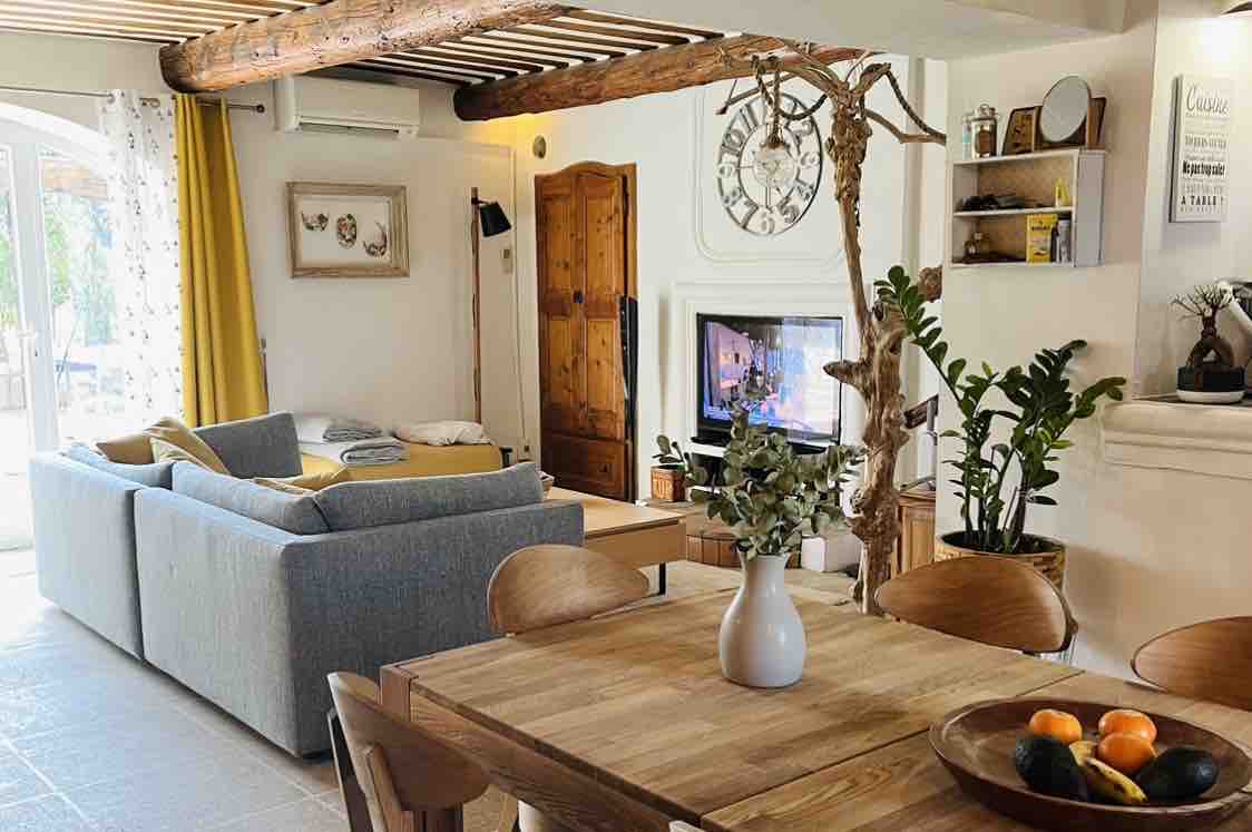 The living and dining area features a spacious gray sofa positioned near a flat-screen television. A wooden dining table with several chairs is complemented by a vase of greenery and fresh fruit. Sunlight filters through large windows, illuminating the warm tones of the interior.