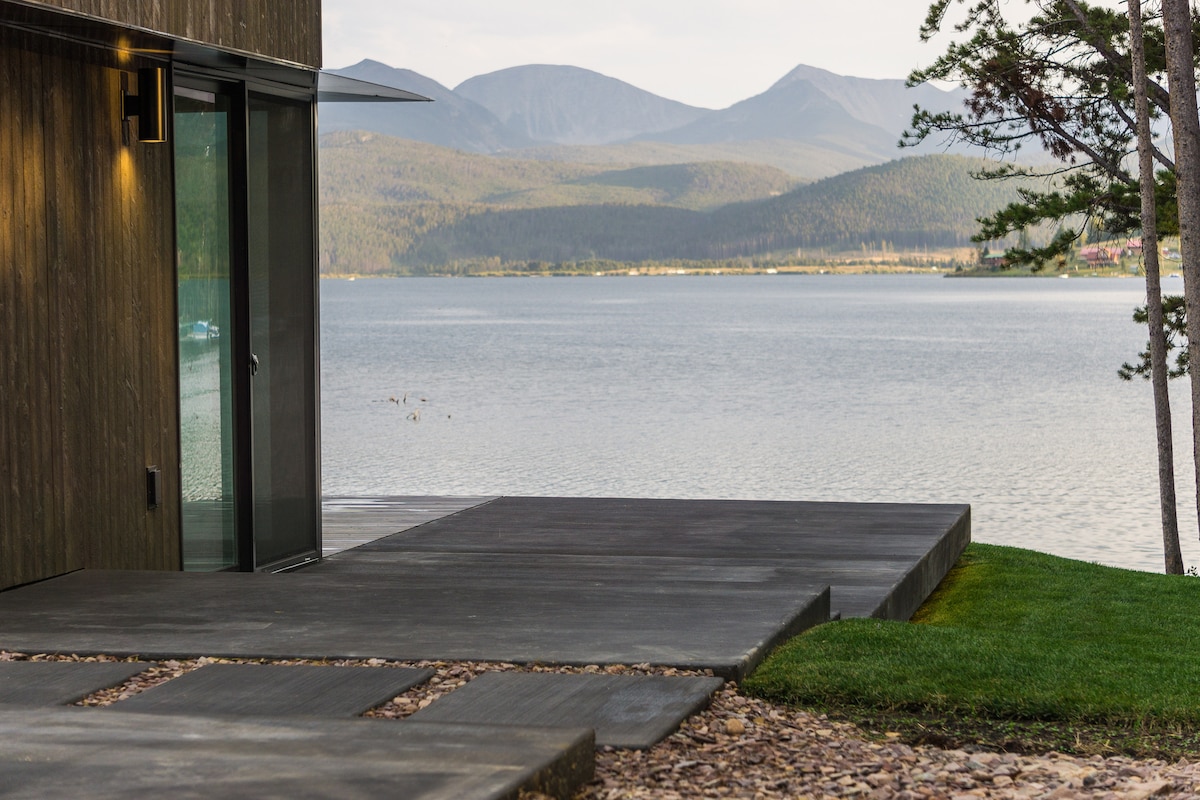 A view of the cabin's exterior reveals a modern stone patio extending toward Georgetown Lake. The serene water reflects the surrounding mountains and a few scattered trees, creating a calm and natural ambiance.