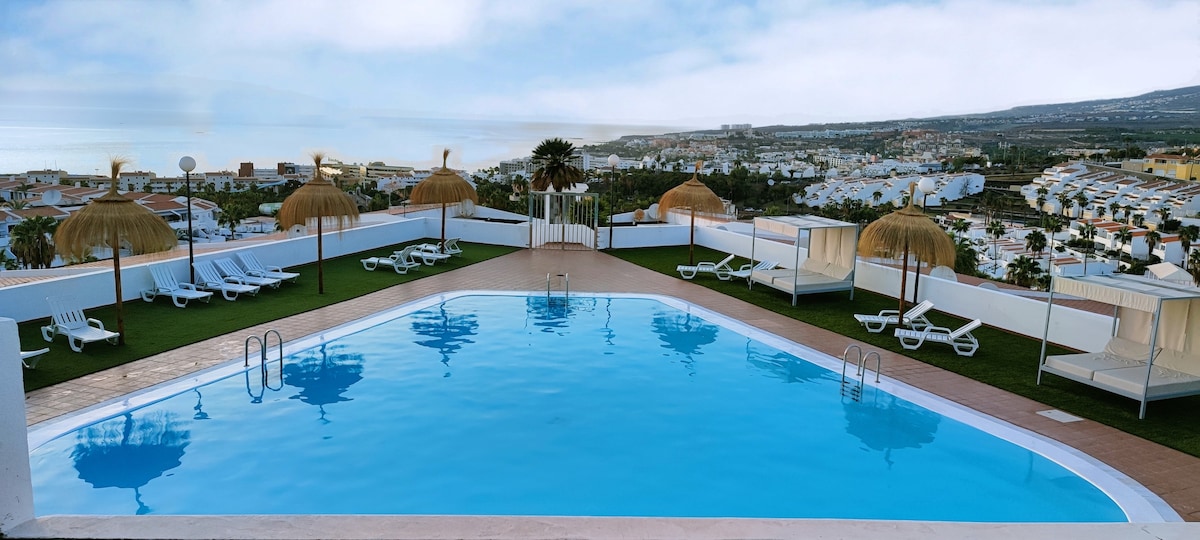A panoramic view of an outdoor pool surrounded by sun loungers and palm-thatched umbrellas. The tranquil water reflects the sky, with the coastline and town visible in the background, offering a sense of openness and relaxation.