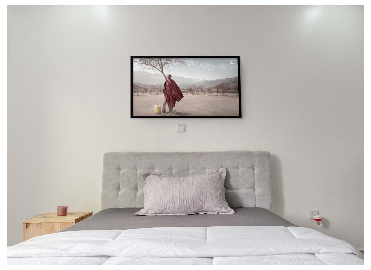 A spacious bed is centered against a clean, neutral wall, topped with a light comforter and a soft decorative pillow. Above the bed, a large framed artwork depicts a serene landscape, featuring a figure in traditional attire amidst natural scenery. Soft lighting enhances the calming ambiance.
