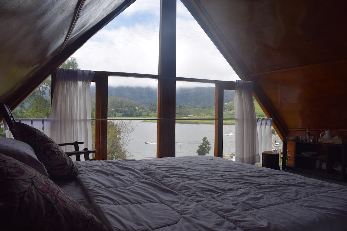 A spacious bedroom is framed by large windows overlooking a serene lake and mountain view. Soft natural light fills the room, highlighting a neatly made bed adorned with patterned pillows. A small seating area with chairs is positioned near the window, enhancing the inviting atmosphere.