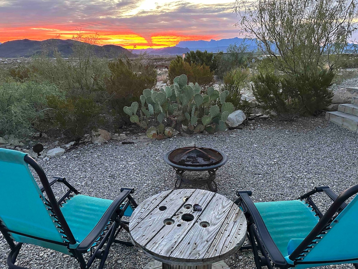 A cozy outdoor setting features two teal reclining chairs placed around a rustic wooden table, positioned near a fire pit. The backdrop showcases a vibrant sunset over distant mountains, with desert vegetation surrounding the area.