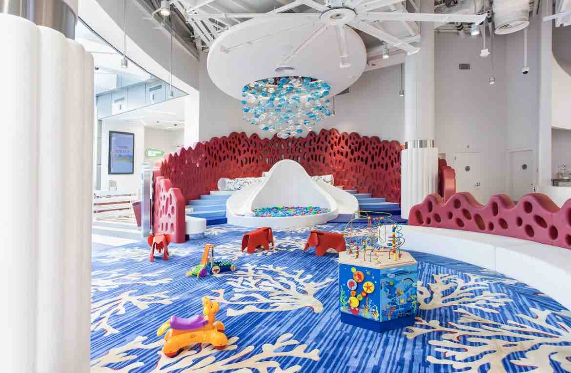 A bright play area features colorful soft seating shaped like a wave, surrounded by coral-inspired walls. An array of toys, including a colorful activity table and small animal figures, is arranged on a vibrant blue carpet adorned with coral designs.