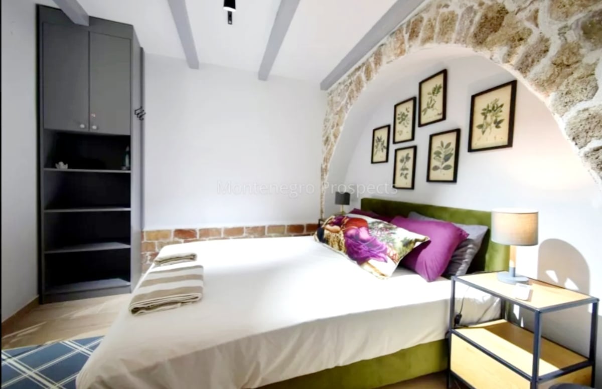 A bright bedroom features a spacious bed adorned with colorful pillows. An accent wall made of natural stone adds texture. Framed botanical prints are hung above the bed, and a small bedside table with a lamp is positioned on one side. A wardrobe is visible in the corner.