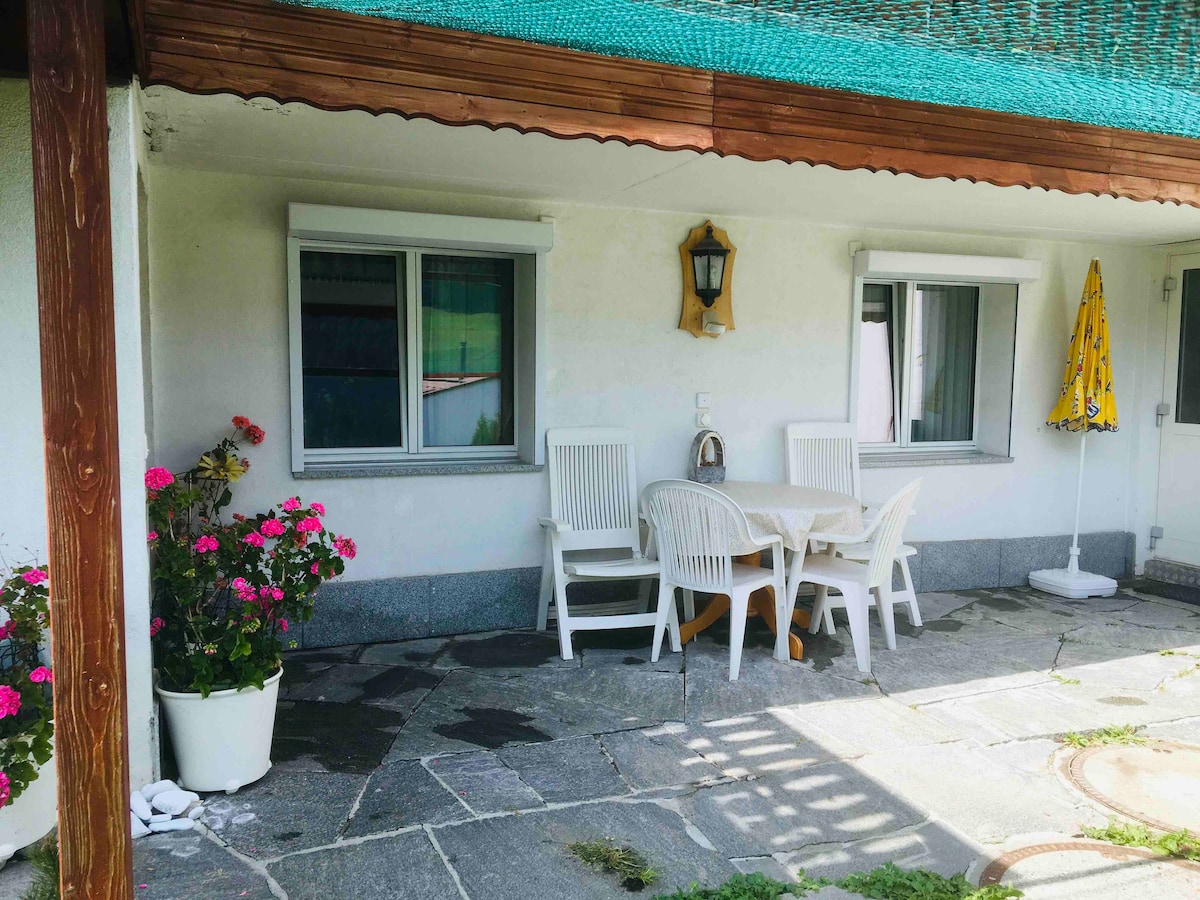A covered patio is presented with a round table and four white chairs, surrounded by flower pots filled with colorful blooms. A sun umbrella stands nearby, while the stone flooring adds a natural touch. Two windows provide a view of the outdoor space.
