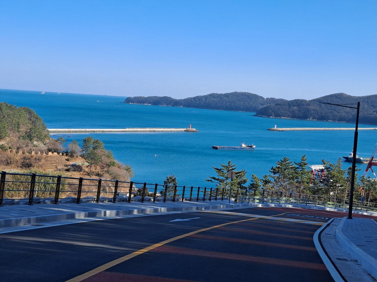 A scenic coastal view is captured, showcasing a vibrant blue sea under a clear sky. The shoreline features a harbor with boats and a breakwater extending into the water. A gently sloped road leads down toward the waterfront, lined with trees and greenery.