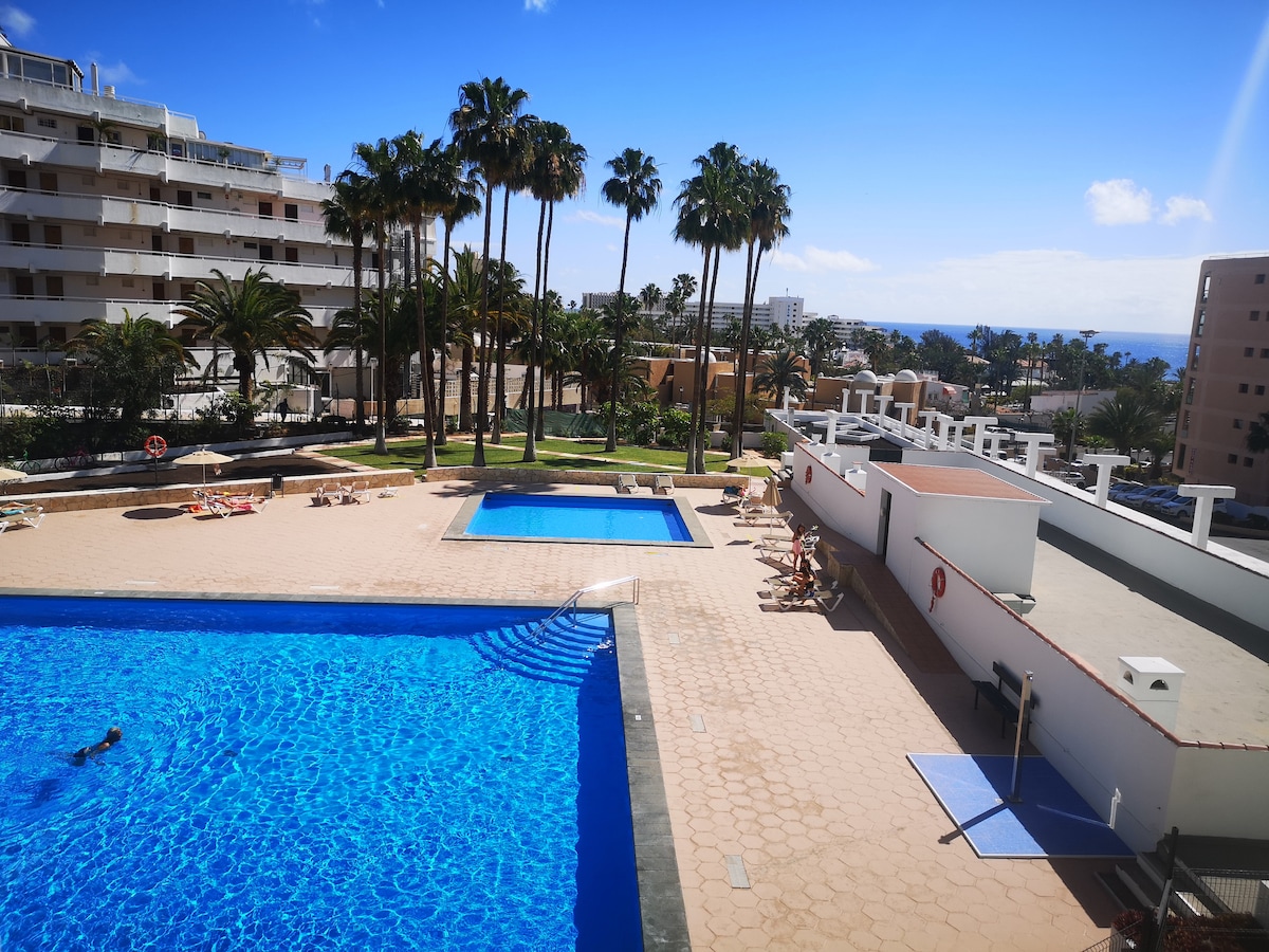 A large salt-water pool is visible, surrounded by a spacious sun terrace. Lush palm trees line the perimeter, providing a tropical feel. A smaller pool for children is situated nearby, with lounge chairs positioned for relaxation. The sea can be seen in the distance under a clear blue sky.